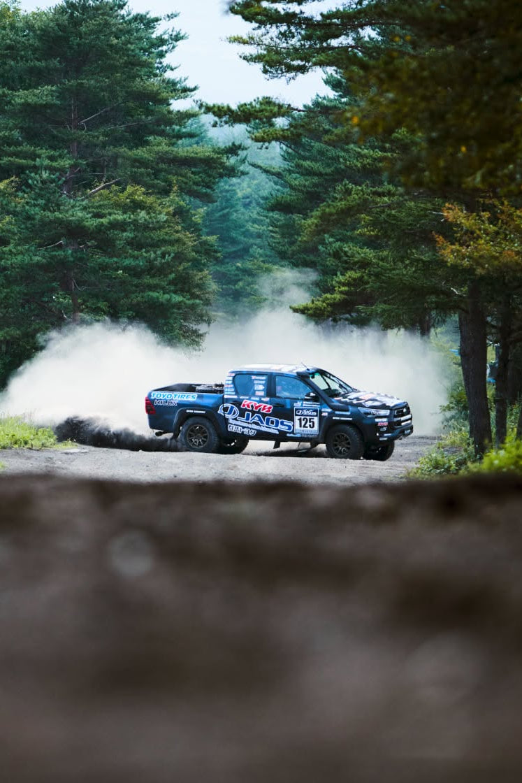 A blue rally truck drifts around a dirt corner on a forest road, kicking up a large cloud of dust. Tall green trees line both sides of the road, and the foreground is blurred.