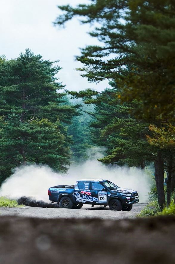 A blue and white rally truck drifts on a dirt road, kicking up a large cloud of dust, with tall green trees in the background.