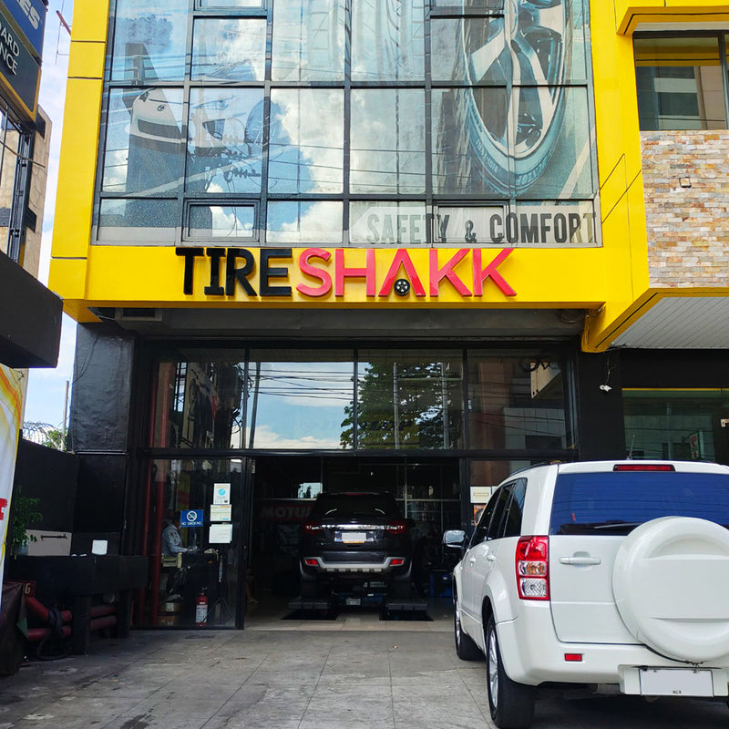 Ortigas Tireshakk auto repair shop storefront with a bright yellow facade, large glass windows, and a white SUV parked in front on a paved street.