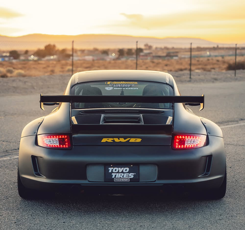 Rear view of a TOYO TIRES matte black Porsche 911 GT3 RWD race car on an asphalt track during sunset, featuring a large rear wing, red taillights, and a Toyo Tires license plate frame.